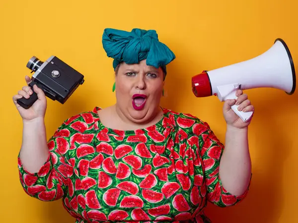 Elinde megafon ve klasik bir kamera olan karpuz elbiseli, etkileyici, iri yarı bir kadın parlak turuncu bir arka plan önünde enerjik bir şekilde bağırıyor..