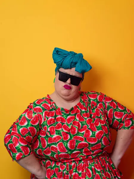 Kendine güvenen, karpuz desenli elbise ve deniz mavisi kafa örtüsü takan, koyu renk güneş gözlüğü takan, turuncu bir arka plana karşı ciddi bir ifadeyle poz veren iri yarı bir kadın..