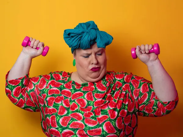Artı renkli karpuz desenli elbise ve deniz mavisi başlıklı kadın turuncu bir arka plana karşı komik ve konsantre bir yüz ifadesiyle pembe halterleri kaldırıyor..
