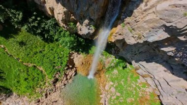 Bereketli yeşil ağaçlar ve ormanlarla çevrili kayalık bir uçurumdan şelalenin havadan görünüşü güneşli bir yaz gününde yumuşak FPV insansız hava aracıyla çekilmiştir, doğa için mükemmeldir.