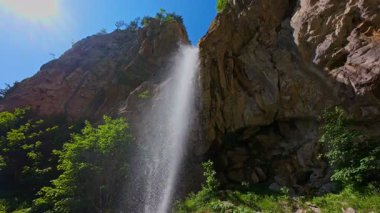 Bereketli yeşil ağaçlar ve ormanlarla çevrili kayalık bir uçurumdan şelalenin havadan görünüşü güneşli bir yaz gününde yumuşak FPV insansız hava aracıyla çekilmiştir, doğa için mükemmeldir.
