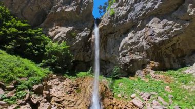 Bereketli yeşil ağaçlar ve ormanlarla çevrili kayalık bir uçurumdan şelalenin havadan görünüşü güneşli bir yaz gününde yumuşak FPV insansız hava aracıyla çekilmiştir, doğa için mükemmeldir.