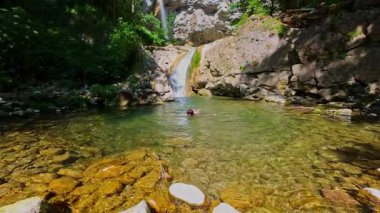 Güneşli bir günde yemyeşil orman ve kayalarla çevrili temiz bir dağ nehrinde yüzen bir kadının FPV drone görüntüsü. Vahşi doğada yaz kaçamağı, macera ve seyahat için ideal.. 