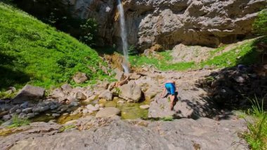 FPV insansız hava aracı görüntüsü, çıplak ayak dengede duran bir adamın manzaralı bir şelalenin, kayalık kayalıkların ve ormandaki yemyeşil bir arazinin yakınındaki bir kütük üzerinde. Yaz macerası, yürüyüş ve doğa keşfi konsepti..