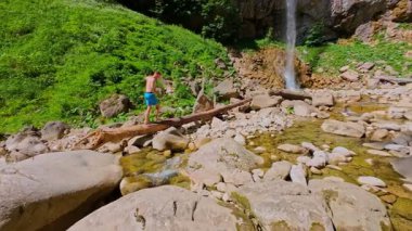 FPV insansız hava aracı görüntüsü, çıplak ayak dengede duran bir adamın manzaralı bir şelalenin, kayalık kayalıkların ve ormandaki yemyeşil bir arazinin yakınındaki bir kütük üzerinde. Yaz macerası, yürüyüş ve doğa keşfi konsepti..