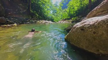 Güneşli bir günde yemyeşil orman ve kayalarla çevrili temiz bir dağ nehrinde yüzen bir kadının FPV drone görüntüsü. Vahşi doğada yaz kaçamağı, macera ve seyahat için ideal.. 