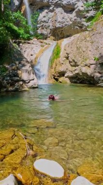 Güneşli bir günde yemyeşil orman ve kayalarla çevrili temiz bir dağ nehrinde yüzen bir kadının FPV drone görüntüsü. Vahşi doğada yaz kaçışı, macera ve seyahat kavramının tazelenmesi 