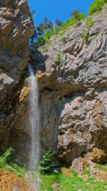 Bereketli yeşil ağaçlar ve ormanlarla çevrili kayalık bir uçurumdan şelalenin havadan çekilmiş dikey videosu güneşli bir yaz gününde FPV insansız hava aracıyla ağır çekimde çekildi..