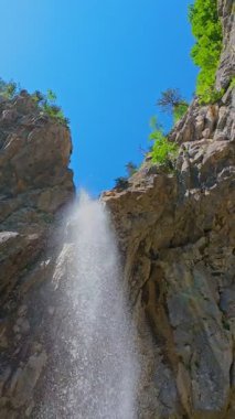 Bereketli yeşil ağaçlar ve ormanlarla çevrili kayalık bir uçurumdan şelalenin havadan çekilmiş dikey videosu güneşli bir yaz gününde FPV insansız hava aracıyla ağır çekimde çekildi..