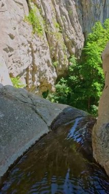 Bereketli yeşil ağaçlar ve ormanlarla çevrili kayalık bir uçurumdan şelalenin havadan çekilmiş dikey videosu güneşli bir yaz gününde FPV insansız hava aracıyla ağır çekimde çekildi..