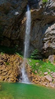 Bereketli yeşil ağaçlar ve ormanlarla çevrili kayalık bir uçurumdan şelalenin havadan çekilmiş dikey videosu güneşli bir yaz gününde FPV insansız hava aracıyla ağır çekimde çekildi..