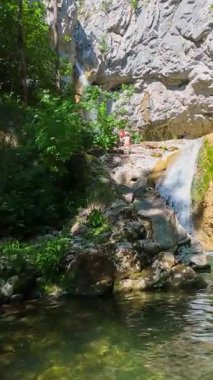 Bereketli yeşil ağaçlar ve ormanlarla çevrili kayalık bir uçurumdan şelalenin havadan çekilmiş dikey videosu güneşli bir yaz gününde FPV insansız hava aracıyla ağır çekimde çekildi..