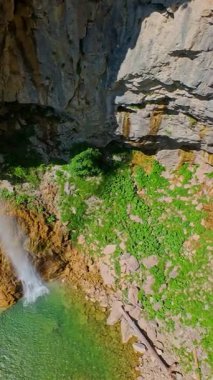 Bereketli yeşil ağaçlar ve ormanlarla çevrili kayalık bir uçurumdan şelalenin havadan çekilmiş dikey videosu güneşli bir yaz gününde FPV insansız hava aracıyla ağır çekimde çekildi..
