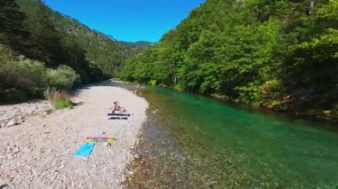 Berrak yeşil bir nehrin havadan görünüşü yemyeşil bir orman ve dağ vadisinde akıyor. İnsanlar canlı bir mavi gökyüzü kaçışı altında bir çakıl taşı plajında dinleniyor.