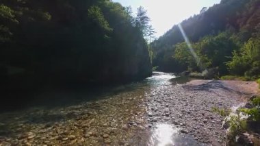 Berrak yeşil bir nehrin havadan görünüşü yemyeşil bir orman ve dağ vadisinde akıyor. İnsanlar canlı bir mavi gökyüzü kaçışı altında bir çakıl taşı plajında dinleniyor.