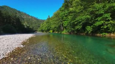 Berrak yeşil bir nehrin havadan görünüşü yemyeşil bir orman ve dağ vadisinde akıyor. İnsanlar canlı bir mavi gökyüzü kaçışı altında bir çakıl taşı plajında dinleniyor.