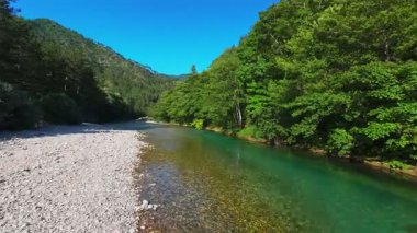 Berrak yeşil bir nehrin havadan görünüşü yemyeşil bir orman ve dağ vadisinde akıyor. İnsanlar canlı bir mavi gökyüzü kaçışı altında bir çakıl taşı plajında dinleniyor.