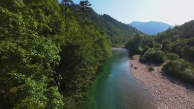 Berrak yeşil bir nehrin havadan görünüşü yemyeşil bir orman ve dağ vadisinde akıyor. İnsanlar canlı bir mavi gökyüzü kaçışı altında bir çakıl taşı plajında dinleniyor.