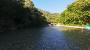 Berrak yeşil bir nehrin havadan görünüşü yemyeşil bir orman ve dağ vadisinde akıyor. İnsanlar canlı bir mavi gökyüzü kaçışı altında bir çakıl taşı plajında dinleniyor.