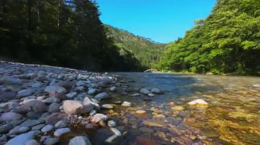 Berrak yeşil bir nehrin havadan görünüşü yemyeşil bir orman ve dağ vadisinde akıyor. İnsanlar canlı bir mavi gökyüzü kaçışı altında bir çakıl taşı plajında dinleniyor.