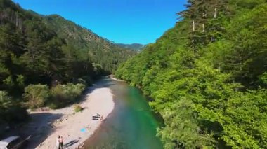 Berrak yeşil bir nehrin havadan görünüşü yemyeşil bir orman ve dağ vadisinde akıyor. İnsanlar canlı bir mavi gökyüzü kaçışı altında bir çakıl taşı plajında dinleniyor.