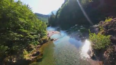 Berrak yeşil bir nehrin havadan görünüşü yemyeşil bir orman ve dağ vadisinde akıyor. İnsanlar canlı bir mavi gökyüzü kaçışı altında bir çakıl taşı plajında dinleniyor.