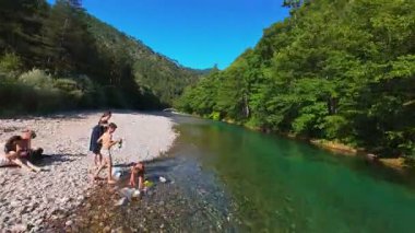 Berrak yeşil bir nehrin havadan görünüşü yemyeşil bir orman ve dağ vadisinde akıyor. İnsanlar canlı bir mavi gökyüzü kaçışı altında bir çakıl taşı plajında dinleniyor.