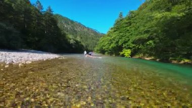 Arkadaşların FPV insansız hava aracı görüntüsü, sığ, kristal berrak bir nehirde, yemyeşil ormanlarla çevrili, parlak mavi gökyüzü altındaki uzak dağlarda, yaz macerasında yüzer.