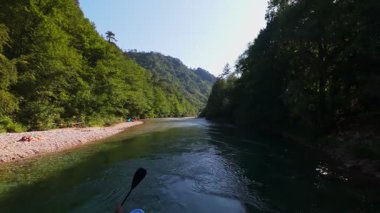 Arkadaşların FPV insansız hava aracı görüntüsü, sığ, kristal berrak bir nehirde, yemyeşil ormanlarla çevrili, parlak mavi gökyüzü altındaki uzak dağlarda, yaz macerasında yüzer.