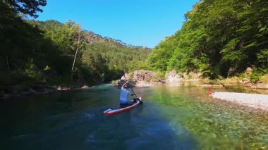 Arkadaşların FPV insansız hava aracı görüntüsü, sığ, kristal berrak bir nehirde, yemyeşil ormanlarla çevrili, parlak mavi gökyüzü altındaki uzak dağlarda, yaz macerasında yüzer.