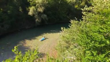 Arkadaşların FPV insansız hava aracı görüntüsü, sığ, kristal berrak bir nehirde, yemyeşil ormanlarla çevrili, parlak mavi gökyüzü altındaki uzak dağlarda, yaz macerasında yüzer.