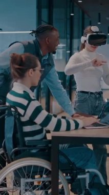 Modern bir iş yerinde çalışan tekerlekli sandalyedeki engelli bir iş kadını. Çevredeki ekip sanal gerçeklik gözlükleri kullanıyor, açık alan yaratıcılığında en son teknolojiyle işbirliği yapıyorlar.