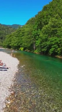 Berrak yeşil bir nehrin havadan görünüşü yemyeşil bir orman ve dağ vadisinde akıyor. İnsanlar canlı bir mavi gökyüzü kaçışı altında bir çakıl taşı plajında dinleniyor.