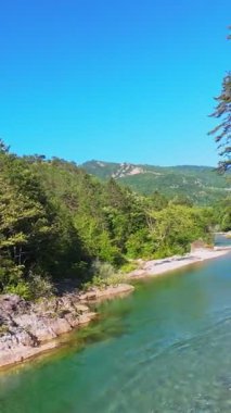 FPV hava aracının dikey videosu. Dostlar kürekleri sığ kristal berrak bir nehirde, yemyeşil orman ve açık mavi gökyüzü altında uzak dağlarla çevrili, yaz macerası ve...