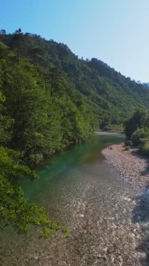 Kristal berraklığında yeşil bir nehrin dikey hava manzarası yemyeşil bir orman ve dağ vadisinde akıyor, insanlar canlı mavi bir yaz gökyüzü altında bir çakıl taşı plajında dinleniyor, doğa kaçışı, sükunet