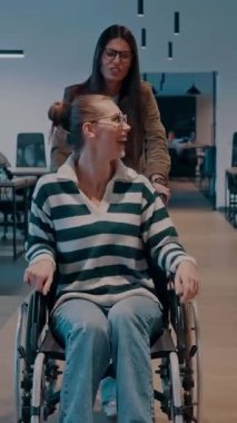 Modern bir ofiste neşeli bir an. Tekerlekli sandalyedeki bir kadın meslektaşı tarafından itilir, ikisi de gülümser. Girişimcilik alanında özel işyeri, takım çalışması, çeşitlilik ve erişilebilirlik