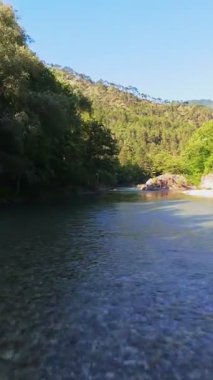Kristal berraklığında yeşil bir nehrin dikey hava manzarası yemyeşil bir orman ve dağ vadisinde akıyor, insanlar canlı mavi bir yaz gökyüzü altında bir çakıl taşı plajında dinleniyor, doğa kaçışı, sükunet