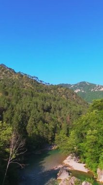 Kristal berraklığında yeşil bir nehrin dikey hava manzarası yemyeşil bir orman ve dağ vadisinde akıyor, insanlar canlı mavi bir yaz gökyüzü altında bir çakıl taşı plajında dinleniyor, doğa kaçışı, sükunet