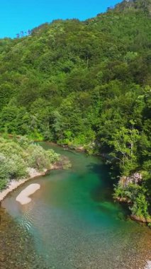 Kristal berraklığında yeşil bir nehrin dikey hava manzarası yemyeşil bir orman ve dağ vadisinde akıyor, insanlar canlı mavi bir yaz gökyüzü altında bir çakıl taşı plajında dinleniyor, doğa kaçışı, sükunet