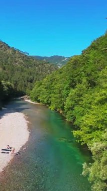 Kristal berraklığında yeşil bir nehrin dikey hava manzarası yemyeşil bir orman ve dağ vadisinde akıyor, insanlar canlı mavi bir yaz gökyüzü altında bir çakıl taşı plajında dinleniyor, doğa kaçışı, sükunet