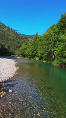 Kristal berraklığında yeşil bir nehrin dikey hava manzarası yemyeşil bir orman ve dağ vadisinde akıyor, insanlar canlı mavi bir yaz gökyüzü altında bir çakıl taşı plajında dinleniyor, doğa kaçışı, sükunet