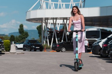 Pembe elbiseli ve güneş gözlüklü zarif bir kadın güneşli şehir sokaklarında elektrikli scooterla geziyor. Mükemmel bir şehir macerasının özgürlüğünü ve tarzını kucaklıyor.