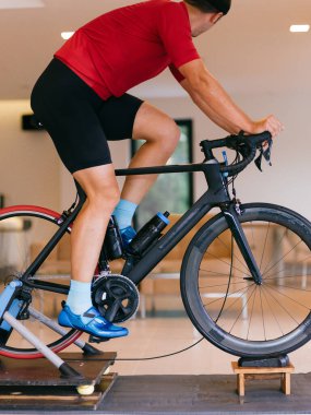 Kapalı mekanda bisiklete binen odaklanmış atletik bir adam kırmızı gömlek ve siyah saç bandı takıyor, yoğun yüz ifadesi takınıyor, evde egzersiz yapıyor..