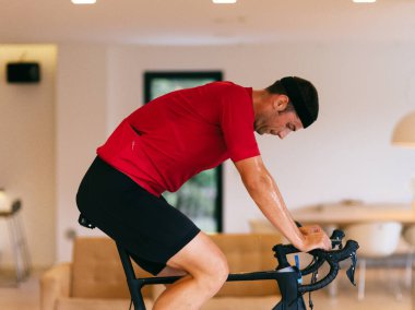 Spor kıyafetleri ve siyah saç bandı takan triatloncunun modern bir evde durgun bir bisiklette bisiklet sürüşünün yan görüntüsü..