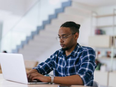 Ekose gömlekli neşeli, Afro-Amerikan bir adam ofis masasında dizüstü bilgisayarla çalışıyor..