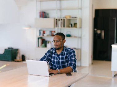 Ekose gömlekli neşeli, Afro-Amerikan bir adam ofis masasında dizüstü bilgisayarla çalışıyor..