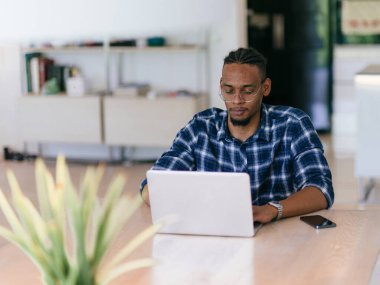 Ekose gömlekli neşeli, Afro-Amerikan bir adam ofis masasında dizüstü bilgisayarla çalışıyor..