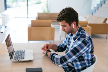 Modern bir oturma odasındaki masada oturan bir serbest çalışan, kulaklıklarıyla iş görüşmesi için bir laptop kullanan, arkadaşlarıyla sohbet eden ve eğlenen biri..