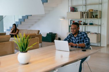 Modern bir oturma odasındaki masada oturan gözlüklü Afrikalı Amerikalı etkileyiciler iş görüşmesi, arkadaşlarla sohbet ve eğlence için dizüstü bilgisayar kullanıyorlar.
