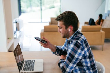 Modern bir oturma odasındaki masada oturan bir serbest çalışan, kulaklıklarıyla iş görüşmesi için bir laptop kullanan, arkadaşlarıyla sohbet eden ve eğlenen biri..